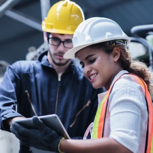 2 employees talking over a work document with hard hats on. one white male with glasses and a beard ib a grey  hoodie and the other a hispanic female with a white shirt and a safety vest