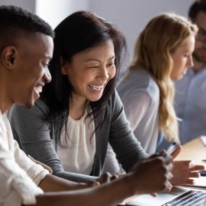 Black mand and asian wonam smiling and talking over a laptop. white blonde woman and white man with glasses talking over a laptop in the background